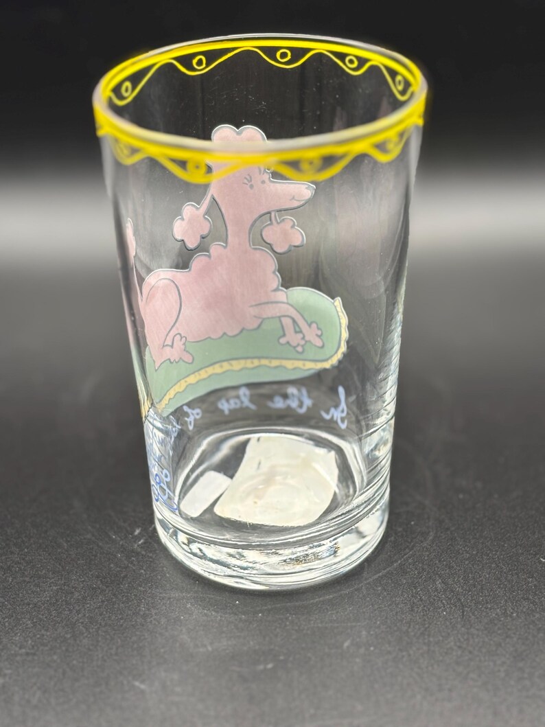 May include: Clear glass tumbler with a pink poodle illustration resting on a green pillow. The rim is decorated with a yellow scalloped design. The glass has text in cursive script near the base.