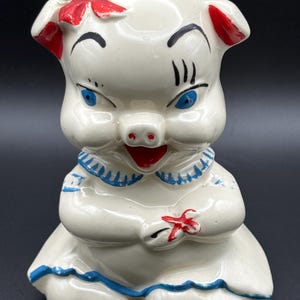 May include: A ceramic piggy bank figurine featuring a white pig with a red bow on its ear, blue eyes, and black eyelashes. The pig wears a white dress with a blue collar and hem, and holds a small red flower in its hands against a dark background.