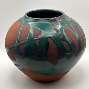 May include: A handcrafted ceramic vase with a rounded shape. The vase features a dark teal glaze over a reddish-brown base, with a pattern of irregular shapes. The interior is dark, and the vase has a wide opening.