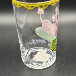 May include: Clear glass with a yellow rim and a pink poodle illustration. The poodle is lounging on a green cushion. The glass has a handwritten signature and the words "great" and "4" in blue and green.