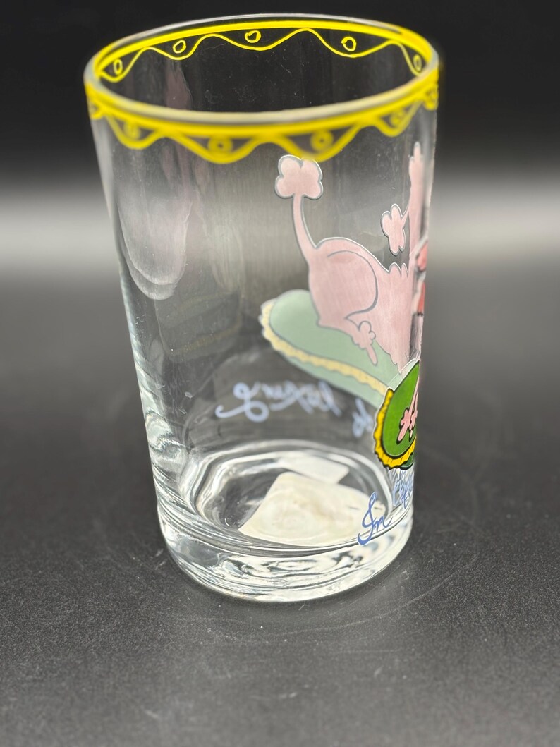 May include: Clear glass with a yellow rim and a pink poodle illustration. The poodle is lounging on a green cushion. The glass has a handwritten signature and the words "great" and "4" in blue and green.