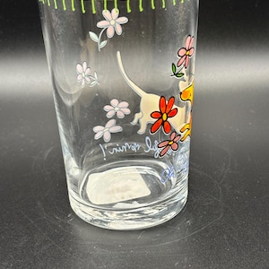 May include: Clear glass tumbler with a green rim and floral and dog illustrations. The glass features a whimsical design with pink and red flowers and a playful dog. The text on the glass reads "junior life".
