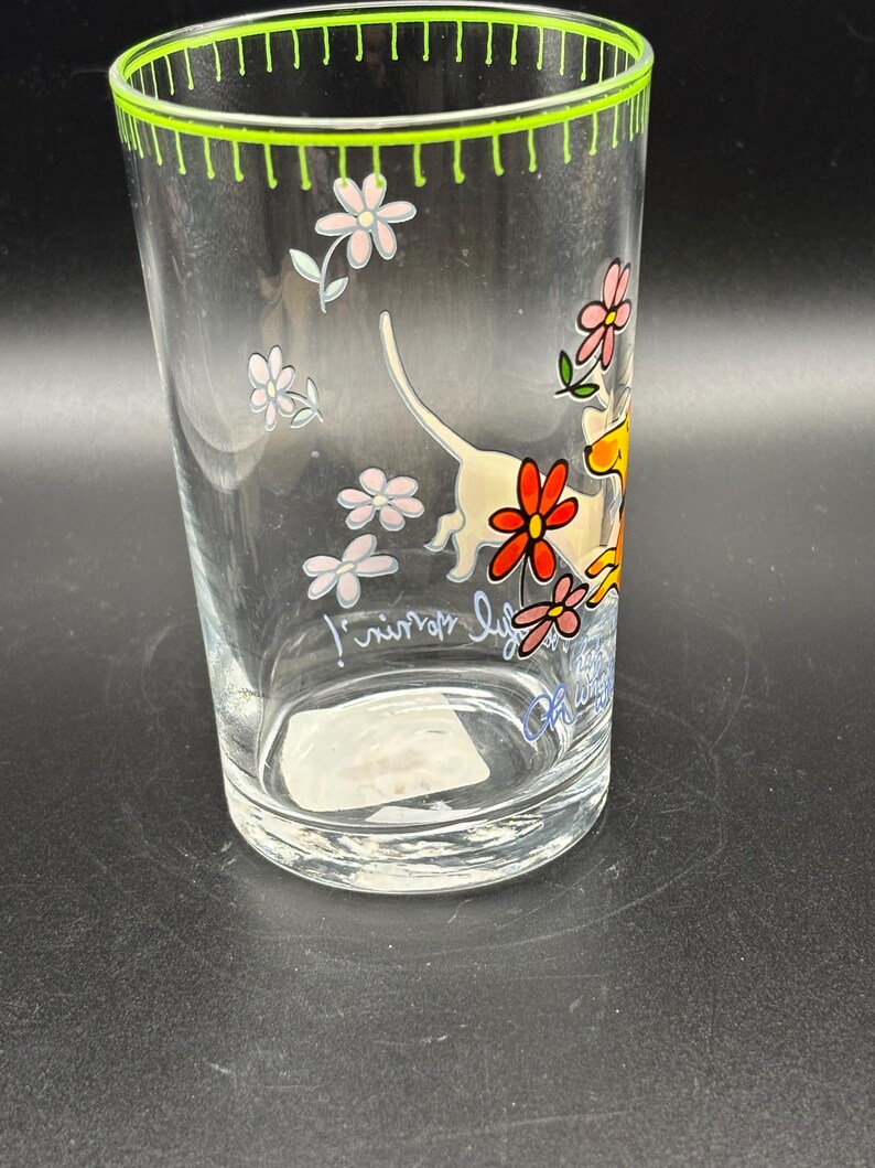 May include: Clear glass tumbler with a green rim and floral and dog illustrations. The glass features a whimsical design with pink and red flowers and a playful dog. The text on the glass reads "junior life".
