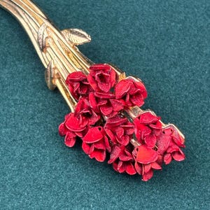 May include: A gold-tone metal brooch shaped like a bouquet of flowers. The piece features a long, slender stem with small leaves at the top, leading into a cluster of vibrant, textured red blossoms that form the head of the floral arrangement.