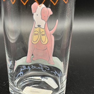May include: Clear glass tumbler with an orange geometric design at the rim. The tumbler features a pink dog illustration with a white face and yellow shoes. The words "fully committed" are written in cursive on the glass.
