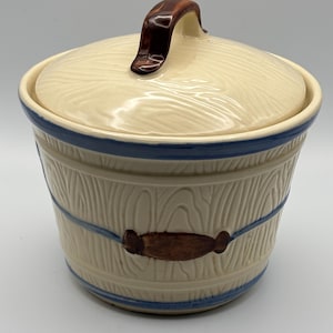 May include: A cream-colored ceramic canister with a lid, featuring a molded wood grain texture. The design includes two horizontal blue stripes and a central brown oval accent. The lid has a brown handle shaped like a small piece of wood.