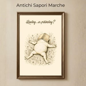 Può includere: Stampa artistica incorniciata con un'illustrazione umoristica di una rana sdraiata nell'erba, con un libro sulla testa. Il testo "Reading...or pretending?" è sopra la rana. La cornice è marrone e lo sfondo è beige neutro.