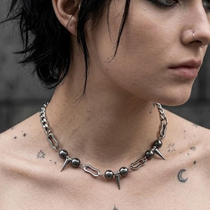 Könnte beinhalten: Silberfarbene Kettenhalskette mit abwechselnden Stachel- und Perlenakzenten. Die Halskette hat ein klobiges Kettendesign mit einer Mischung aus glatten und spitzen Elementen. Die Halskette ist ein auffälliger Schmuck.