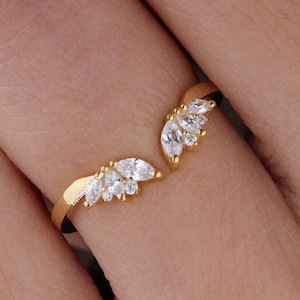 Peut inclure: Une bague dorée avec un design fendu, ornée de grappes de pierres précieuses claires, de taille marquise. La conception ouverte de la bague s'enroule autour du doigt, créant un look unique. Les pierres sont disposées en éventail.