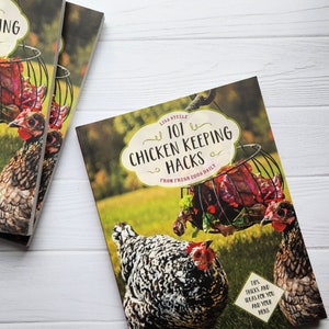 May include: A book titled "101 Chicken Keeping Hacks: From Fresh Eggs Daily" with a colorful cover featuring two chickens and a basket of red lettuce. The book cover has a white background with green grass in the background.