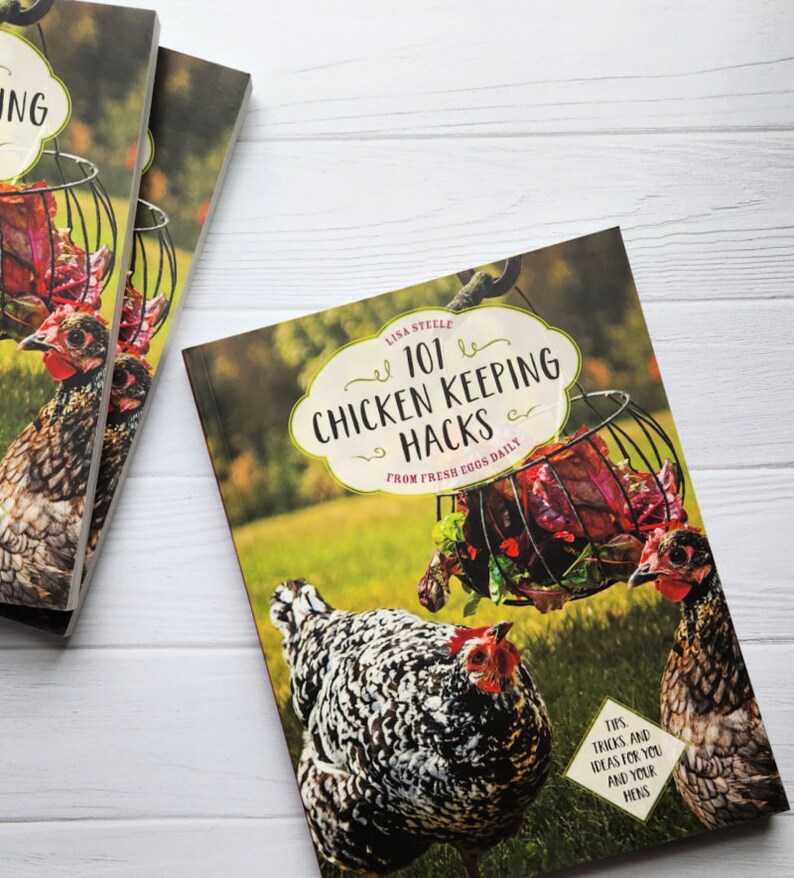 May include: A book titled "101 Chicken Keeping Hacks: From Fresh Eggs Daily" with a colorful cover featuring two chickens and a basket of red lettuce. The book cover has a white background with green grass in the background.