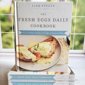 May include: A stack of cookbooks titled "The Fresh Eggs Daily Cookbook" by Lisa Steele. The cover features a photo of eggs benedict on toast. The books are stacked in front of a window with a blurred outdoor view.