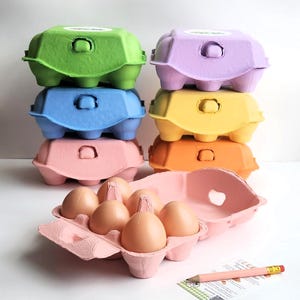 May include: A stack of six colorful cardboard egg cartons in green, lavender, blue, yellow, orange, and pink. One open pink carton holds six brown eggs. A small rectangular card and a yellow pencil with a pink eraser lie next to the open carton.