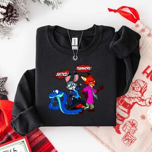 May include: Black crewneck sweatshirt featuring a cartoon illustration of a blue snake, a gray bunny, and a red fox. The words "JUSTICE!" and "TEAMWORK!" are in speech bubbles. A silver necklace is visible.