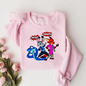 May include: A light pink sweatshirt featuring a cartoon graphic of a blue snake, a gray rabbit, and a red fox. The words "JUSTICE!" and "TEAMWORK!" are in speech bubbles. The sweatshirt has long sleeves.