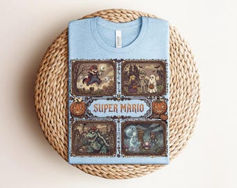 Könnte beinhalten: Ein hellblaues T-Shirt mit einem Grafikdruck, der vier gerahmte Szenen von Super Mario-Charakteren in Halloween-Kostümen zeigt. Der zentrale Text lautet Super Mario, umgeben von dekorativen Rändern, Kürbissen und Totenköpfen im düsteren Stil.