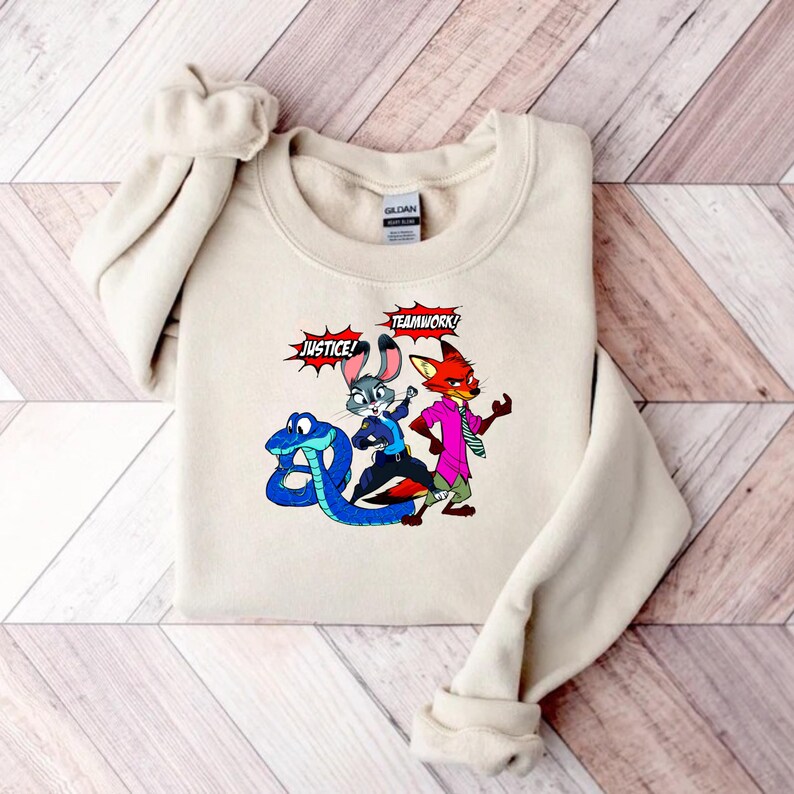 May include: Beige crewneck sweatshirt featuring a cartoon graphic of a blue snake, a gray bunny, and a red fox. The words "JUSTICE!" and "TEAMWORK!" are in speech bubbles. The sweatshirt is made of a soft material.