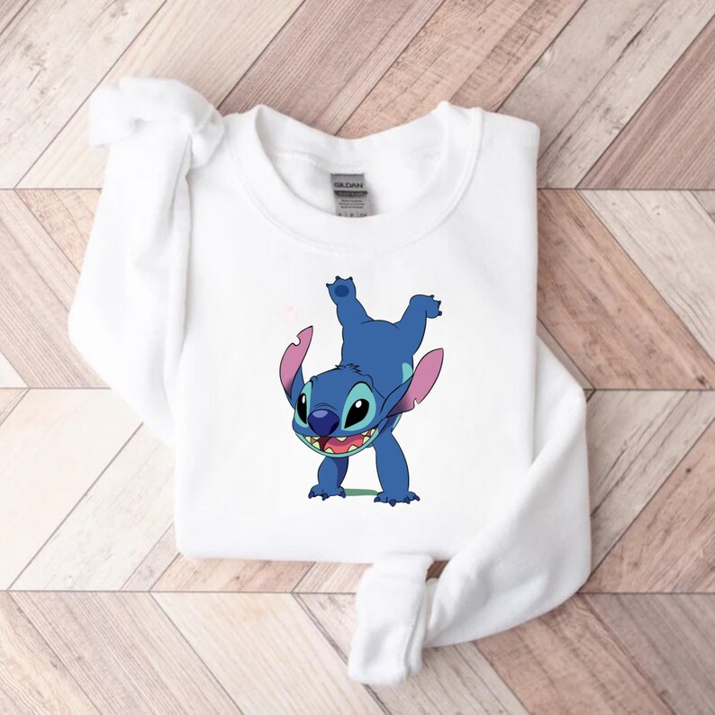 May include: White sweatshirt featuring a blue cartoon character with large ears and a wide grin. The character is in a playful pose, with its arms and legs in the air. The sweatshirt is made of a soft material and has a crew neck.