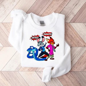 May include: White sweatshirt featuring a colorful cartoon graphic of characters from Zootopia. The design includes a blue snake, a gray rabbit, and a red fox, with the words "JUSTICE!" and "TEAMWORK!" in speech bubbles.