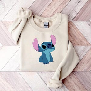 Puede incluir: Una sudadera de color crema con un personaje de dibujos animados azul con grandes orejas y una expresi&oacute;n curiosa. El personaje est&aacute; sentado, con los brazos ligeramente levantados. La sudadera tiene mangas largas y cuello redondo.