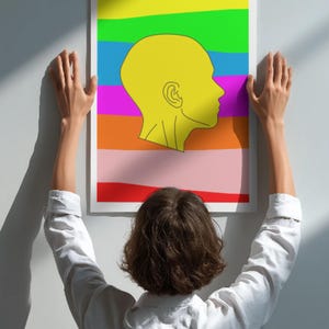 May include: A framed art print featuring a yellow profile against a backdrop of horizontal rainbow stripes. The artwork is being held up by a person wearing a white shirt. The color palette includes red, orange, pink, purple, green, and yellow.