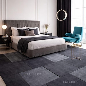 Puede incluir: Un dormitorio moderno con una gran cama gris tapizada con almohadas y una manta gris oscura. Una alfombra con estampado geométrico en tonos grises cubre el suelo. Un sillón turquesa y una mesa auxiliar dorada.