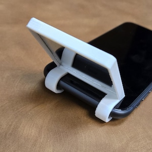 Puede incluir: Un soporte para teléfono blanco impreso en 3D que sostiene un teléfono inteligente negro. El soporte tiene una parte superior rectangular y soportes curvos que envuelven el teléfono. El soporte está diseñado para sostener el teléfono en posición vertical.