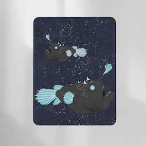 May include: A dark blue rectangular object featuring two anglerfish illustrations. The fish are black with light blue fins and glowing eyes, set against a starry background. The design has a whimsical, aquatic theme.
