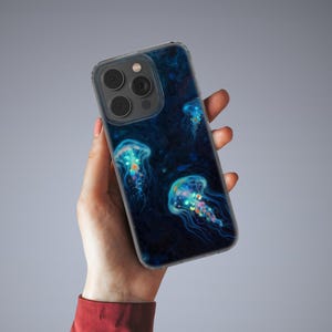 Puede incluir: Una funda transparente para teléfono con un diseño azul oscuro que presenta ilustraciones de medusas brillantes. Las medusas tienen acentos azules, amarillos y naranjas. La funda del teléfono se sostiene en una mano.