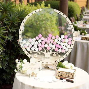 Puede incluir: Un libro de visitas de boda redondo de cristal transparente con un marco de madera cortado con láser. El diseño presenta un árbol con recortes de flores rosas y blancas. Incluye un soporte de madera, una caja pequeña y bolígrafos.