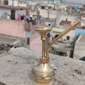 May include: A small, ornate brass hookah with a curved stem and detailed engravings. The hookah is set against a blurred urban backdrop, showcasing its intricate design and golden color. The hookah is approximately 6 inches tall.
