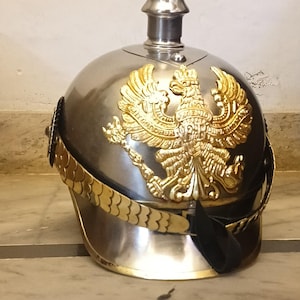 May include: A silver-coloured metal helmet with a tall, pointed top. The helmet features a gold-coloured eagle emblem on the front and a gold-coloured band around the base. A black strap is attached to the helmet.