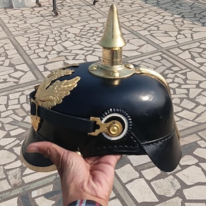 May include: Black Prussian Pickelhaube helmet with gold-colored accents. The helmet features a gold spike, decorative gold eagle emblems, and a chin strap. The helmet is held up against a background of a tiled surface and a cityscape.