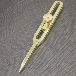 May include: A brass-colored marking gauge tool with a pointed end and an adjustable wheel. The tool has a ruler etched along the side and is designed for precise measurements. It is a woodworking or metalworking tool.