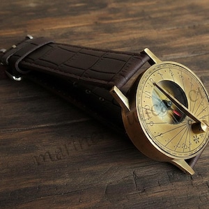 May include: A vintage-style watch with a compass design. The watch features a gold-colored face with intricate markings and a black needle. It has a brown leather band. The watch is set against a wooden surface.