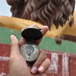 Puede incluir: Una brújula de latón vintage con tapa abierta, sostenida frente a una gran estatua de león. La brújula tiene una esfera detallada con números y marcas. La estatua del león es amarilla y marrón.