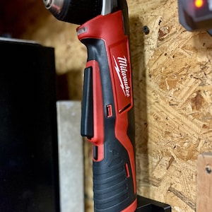 May include: A red and black Milwaukee right-angle drill mounted on a black holder. The drill has a black chuck and a red body with the Milwaukee logo. It is attached to a wooden surface.