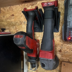 May include: Two red and black power tools, one labeled "FUEL BRUSHLESS", are mounted on a wooden wall. The tools have black handles and red accents. A black charger is visible on the right side of the image.