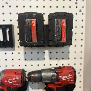 May include: Two black Milwaukee 5.0 Ah battery packs mounted on a white pegboard, positioned above two red and black Milwaukee Fuel power drills. The battery labels feature red and white branding with warning text and technical specifications.