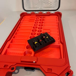 Puede incluir: Maleta de herramientas roja abierta con tapa transparente. En el interior, un soporte negro contiene varias brocas con detalles amarillos y rojos. El logotipo de Milwaukee es visible.