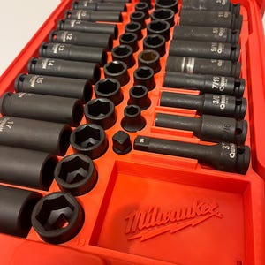 May include: A red toolbox with a variety of black impact sockets and extensions. The sockets are arranged in rows and labeled with sizes in inches. The Milwaukee logo is visible on the case.