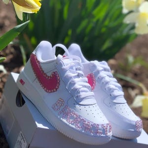 Puede incluir: Zapatillas blancas con detalles rosas enjoyados. El logo de Nike y la palabra "BABY" est&aacute;n adornados con pedrer&iacute;a rosa. Los zapatos est&aacute;n sobre una caja blanca, con un fondo de follaje verde y flores amarillas.