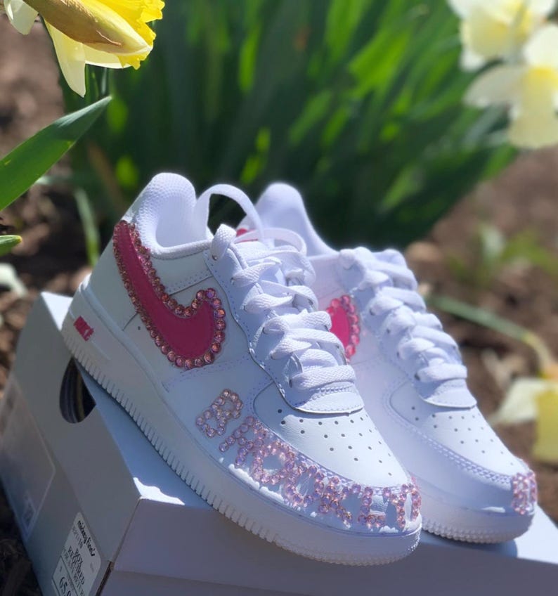 Puede incluir: Zapatillas blancas con detalles rosas enjoyados. El logo de Nike y la palabra "BABY" est&aacute;n adornados con pedrer&iacute;a rosa. Los zapatos est&aacute;n sobre una caja blanca, con un fondo de follaje verde y flores amarillas.