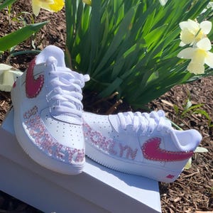 Puede incluir: Zapatillas blancas con detalles rosas, incluyendo un logo Nike rosa y el nombre "Brooklyn" en pedrer&iacute;a rosa. Las zapatillas est&aacute;n sobre una caja blanca, con flores en el fondo.