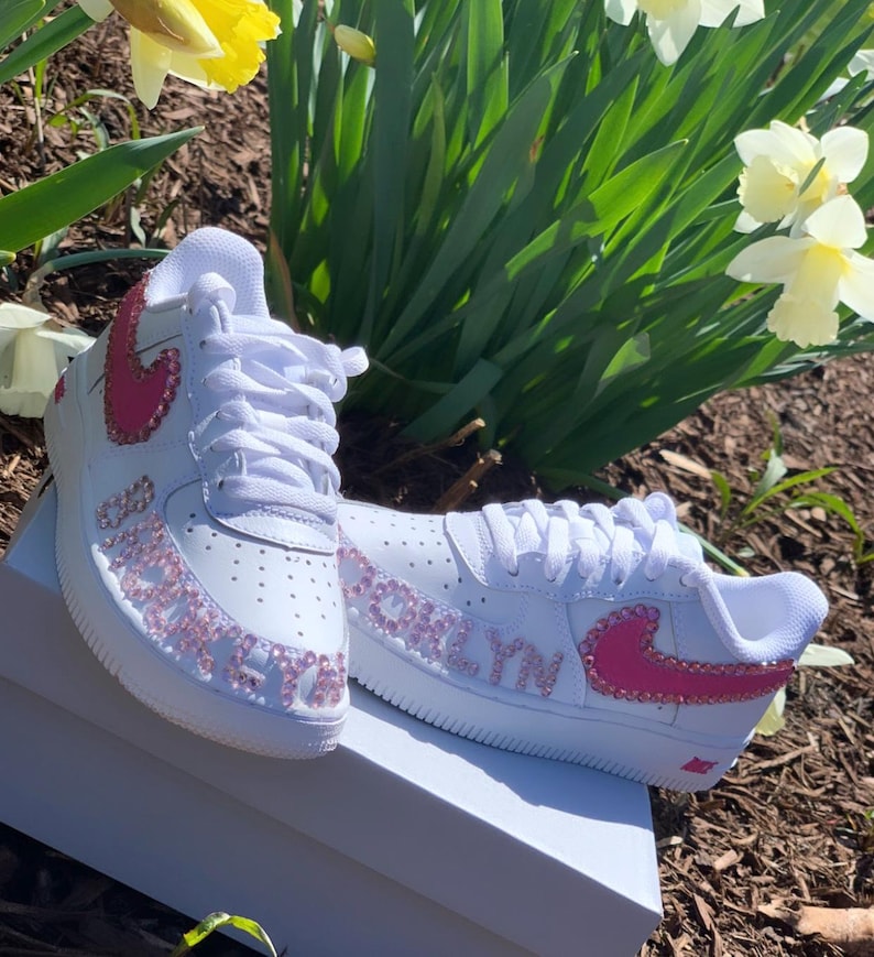Puede incluir: Zapatillas blancas con detalles rosas, incluyendo un logo Nike rosa y el nombre "Brooklyn" en pedrer&iacute;a rosa. Las zapatillas est&aacute;n sobre una caja blanca, con flores en el fondo.