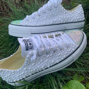 Puede incluir: Zapatillas Converse blancas adornadas con perlas de imitación. Las punteras están decoradas con pedrería iridiscente. Las zapatillas tienen cordones blancos y ribetes negros en las suelas. Se muestran sobre hierba verde.