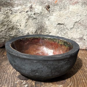 May include: A small, round bowl with a matte, dark charcoal-grey exterior featuring a rough, textured surface. The interior of the bowl has a smooth, glossy finish with mottled shades of copper, rust-orange, and hints of metallic green.
