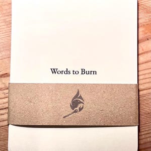 May include: A stack of white rectangular cards with rounded corners, held together by a brown paper band. The text "Words to Burn" is printed in black. The band features a flame and match graphic.