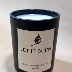 May include: A black candle in a cylindrical glass container. The candle has a white wax interior. A label on the front reads "LET IT BURN" with a graphic of a flame and match. The label also lists the scents: Amber, Patchouli, Clove & Vanilla.