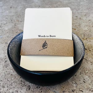 May include: A stack of off-white rectangular cards with the text Words to Burn printed on the top card. The stack is secured by a brown paper band featuring a black flame icon and is placed inside a round bowl with a black and grey speckled finish.